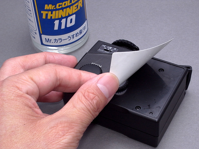 <b>まずは本体のネジを外したいのだが、3個のネジが背面シールの下に隠れている。シールのノリをシンナーで溶かしながら慎重にはがしてゆく</b>