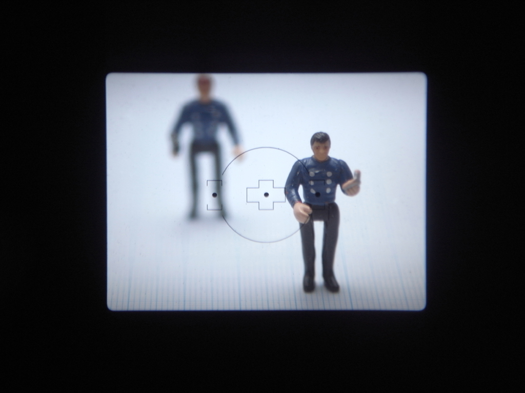 <b>E-420の接眼部からファインダー像を撮影し、交換前後のスクリーンの見え具合の簡単な比較をしてみた。レンズはシグマ「Macro 90mm F2.8」（OMマウント用をアダプターを介して装着）で絞り開放。フィギュアの高さは3cmで手前と奥間隔は5cmである。まずは交換前のE-420に標準装備のスクリーン像。AF主体で使われることを前提に設計されており、明るく見える反面、MF時にピントの山がつかみづらい。また、AF用スーパーインポーズの黒点が、MF時には非常に目障りになる</b>