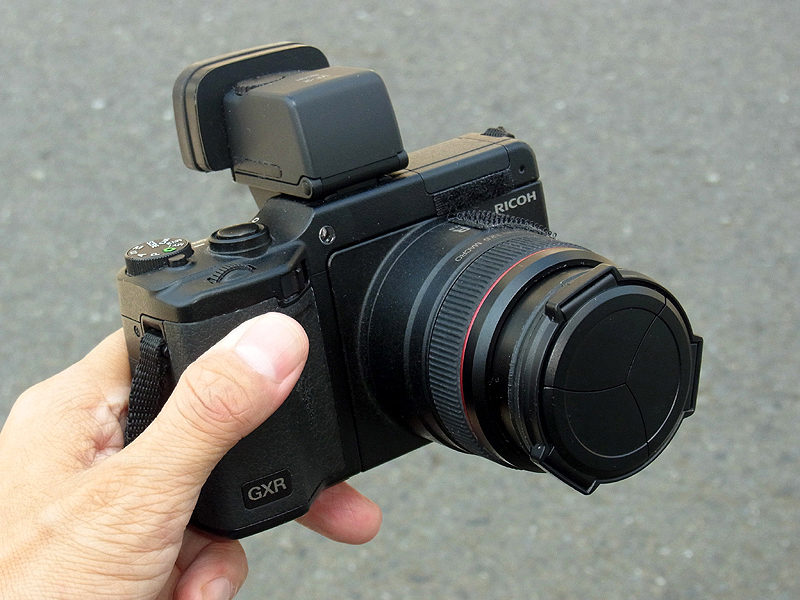 <b>スナップ用カメラとしてさらに進化させた、ぼくのリコーGXR A12ユニット。取り外し式レンズキャップのかわりに、レンズ先端に「自動開閉キャップLC-2」を固定するカスタマイズを施した。<a href="http://dc.watch.impress.co.jp/docs/review/labo/20100705_377760.html"><u>前々回で紹介した</u></a>、倍率アップのカスタムを施した電子ビューファインダー「VF2」も装着している</b>