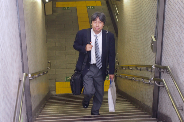 <b>勤務先へは地下鉄で通勤。月曜〜金曜が一般的だが田中さんの部署は行事などによって土日に仕事の場合も少なくない。いつでもバッグには分厚い手帳やパソコンなどがギッシリと詰め込まれていてパンパンに膨らんでいる。そして会社のロゴが入った丈夫な紙袋も資料でいっぱい、肩からはカメラも</b>