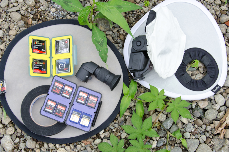 <b>コンパクトストロボとSDHCメモリーカードなどカメラによって違うタイプのメモリカード類がごっそりと。光を拡散して柔らかい発光をさせる、取り外しが容易なデュフューザーを付けたクリップオンストロボ。真っ直ぐな体勢が難しい場合などに有利な、角度を変えて覗けるアングルファインダー。ちょっとした貼り付けなど、何かと便利なパーマセルテープ。2種類の小型デュフューズレフ板など</b>