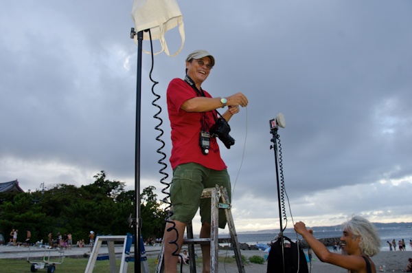 <b>大勢が集まる撮影なので、ハシゴの上から狙う。そして夕暮れ時の逆光でもあり、充電式のストロボの登場だ。太陽の沈む速度と準備との戦いだが仲間みんなで手伝って準備はあっという間に完了。だってみんな熟練したカメラマンばかりだ。太陽さんにも負けるわけがない。ボクのカメラバッグもスタンドの重しとして活躍。ブルースさんの三脚は自転車のベル付きだが、ジッツオでセット販売しているわけではなく、自家製なのでご注意（笑）</b>