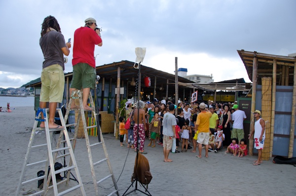 <b>大勢が集まる撮影なので、ハシゴの上から狙う。そして夕暮れ時の逆光でもあり、充電式のストロボの登場だ。太陽の沈む速度と準備との戦いだが仲間みんなで手伝って準備はあっという間に完了。だってみんな熟練したカメラマンばかりだ。太陽さんにも負けるわけがない。ボクのカメラバッグもスタンドの重しとして活躍。ブルースさんの三脚は自転車のベル付きだが、ジッツオでセット販売しているわけではなく、自家製なのでご注意（笑）</b>