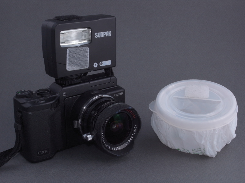 <b>さらにストロボシステムを考えてみる。内蔵ストロボはケラレてしまうため、外部ストロボの「サンパックPF20XD」を装着。ディフューザーはタッパーとコンビニ袋を素材に自作したもので、<a href="http://dc.watch.impress.co.jp/docs/review/labo/20110606_450161.html"><u>「ペンタックス「645D」で試す“ヘビー&amp;ライト”マクロシステム」</u></a>で使用したものと基本的に同一。しかし今回さらに改良を加えている</b>