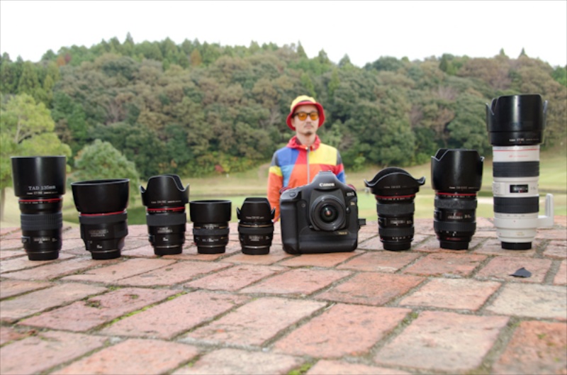 <b>この日のロケにHALくんチームが使った機材を並べてみた。基本的にボディはEOS-1Dsだが、この他に予備としてEOS 5D Mark IIも用意している。向かって左から、EF 135mm F2 L USM、EF 85mm F1.2 L II USM、EF 35mm F1.4 L USM、EF 50mm F1.4 USM、EF 28mm F1.8 USM、EF 24mm F2.8を装着したEOS 1Ds、EF 16-35mm F2.8 L II USM、EF 24-70mm F2.8 L USM、EF 70-200mm F2.8 L USM。後ろにHALくんが立っているのは、階段上で撮影しているので万が一の時に管理責任者として会社所有のレンズが転げ落ちたりしないように構えているから。もう一つの理由は目立ちたいからでもある（笑）</b>