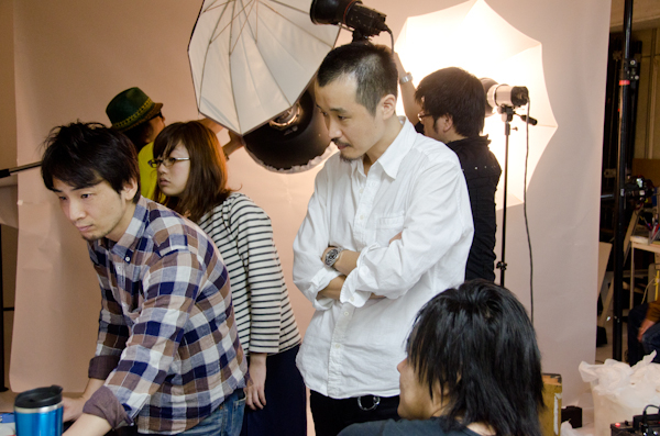 <b>11月中旬の某日。先日のカタログの商品カット撮影のスタジオバージョン。前回のロケでお世話になった皆さんが作業中。横のセットでは野口カメラマンチームは横位置からのバッグを撮影中。どちらも撮影済みカットからレイアウトに当て込んでいくので作業が早くて便利だ。この日のHALくんの担当は商品床置きにしてのカメラ真俯瞰からの撮影。ジッツオの大型三脚にEOS-1Dsを取り付けて、2mくらいの位置からセンターを狙う。キヤノンのユーティリティソフトでパソコンに直結し、パソコン用ディスプレイの大きな画面で確認しながら位置決めをしていく。ファインダーからも直接覗けるようにアングルファインダーを取り付けてある。ボクがお邪魔している間はレンズ交換がなく、24-70mmだけを使用</b>