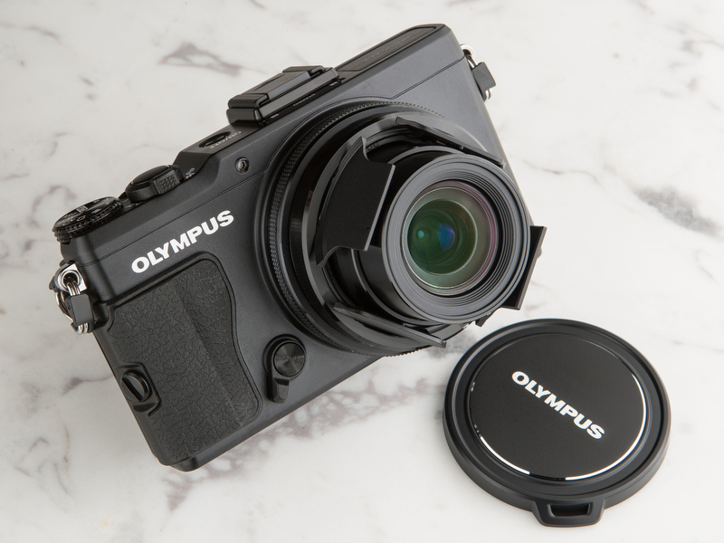 <b>自動開閉レンズキャップを付け、電源投入したところ。右は標準付属のレンズキャップだ。電源オフにすると自動的に閉じる。キャップを着脱する手間が省ける（写真では斜めに装着されているが、カメラ底面に対して縦横のラインが直交するよう、まっすぐ装着できる）</b>