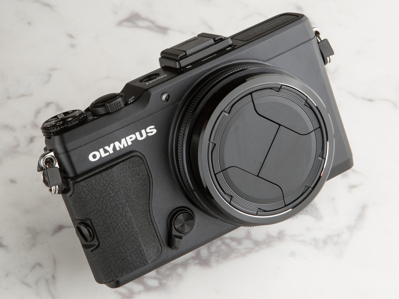 <b>自動開閉レンズキャップを付け、電源投入したところ。右は標準付属のレンズキャップだ。電源オフにすると自動的に閉じる。キャップを着脱する手間が省ける（写真では斜めに装着されているが、カメラ底面に対して縦横のラインが直交するよう、まっすぐ装着できる）</b>