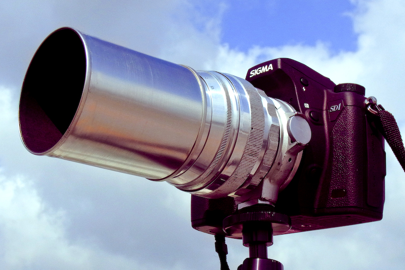 SIGMA SD1 Merrillに装着した、異様にギラギラ輝く銀色のレンズ。旧ソ連製の「Helios（ヘリオス）85mm F1.5」だが、自作の金属製フードを装着し迫力が一層増している。まるで純正品のようにフィットしたこのフード、100円ショップで買った桃缶が素材とはとても思えない（笑）。