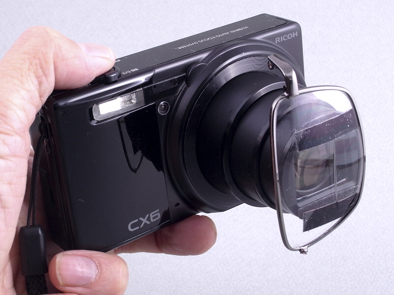 100円ショップで売られていた「二重レンズ老眼鏡」をレンズに装着したリコー「CX6」。これによって1つの画面内に近距離と遠距離の両方にピントのあった写真が撮影できる、特殊カメラに変身した。以前この連載で、ライカレンズに倣って<a class="" href="http://dc.watch.impress.co.jp/docs/review/labo/20110822_467453.html">「FinePix X100」を“メガネ付きファインダー”仕様に改造</a>という記事を書いたのだが。今回のカメラの方が正真正銘“メガネ付き”と言えるだろう(笑)。