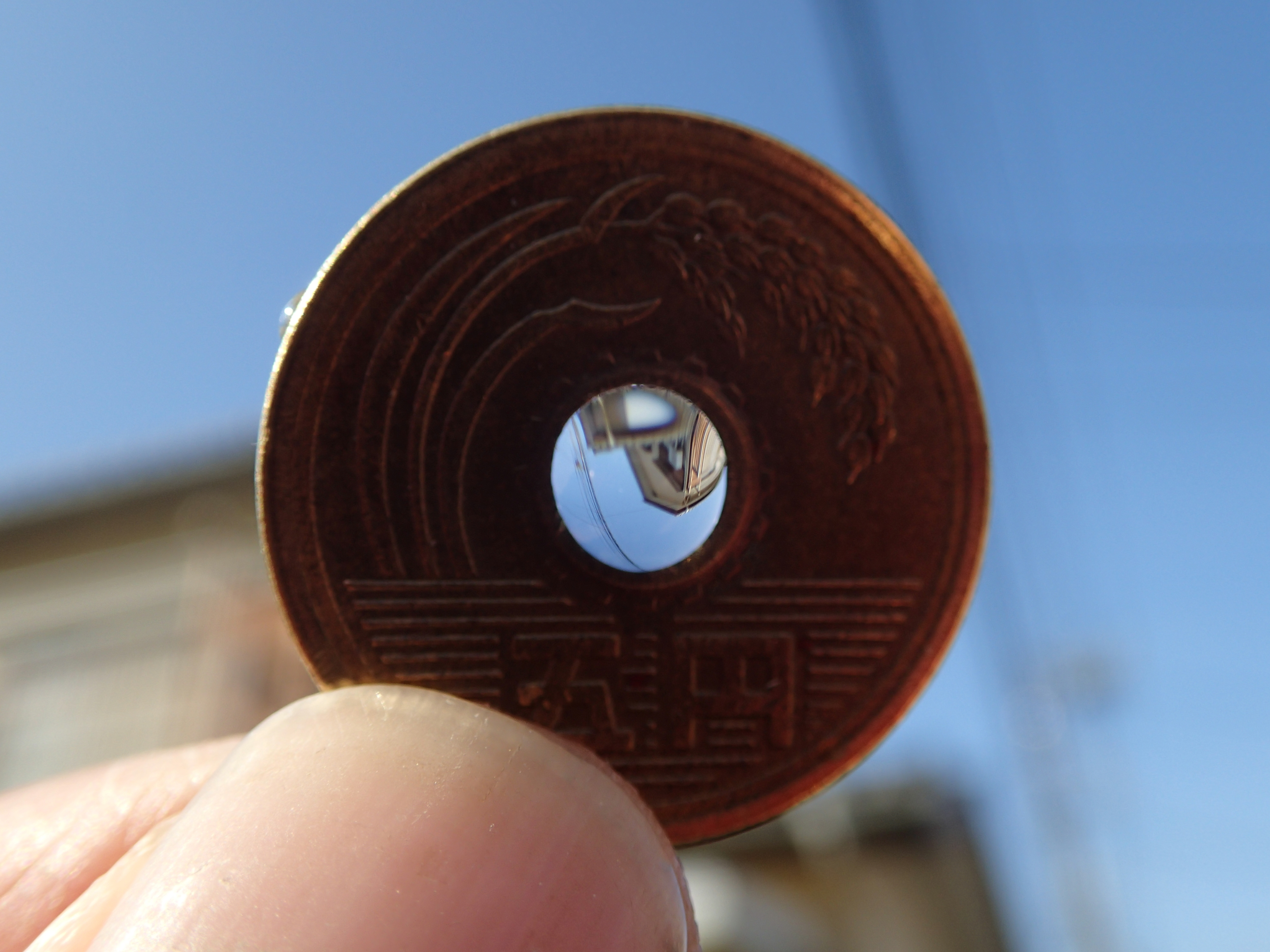 OLYMPUS TG-2スーパーマクロモードの広角側で5円玉レンズを撮影するとこのように写る。指でつまんだ5円玉の真ん中に、家と電線が逆さに写っている。水滴が重力で歪み、映像も歪んで写っているが、表現としてなかなか面白い。レンズ周囲に稲穂の図案があしらわれて、まさに「ミノルタ」と言いたいところだが、残念ながらそのブランドはカメラ業界から無くなってしまった……。