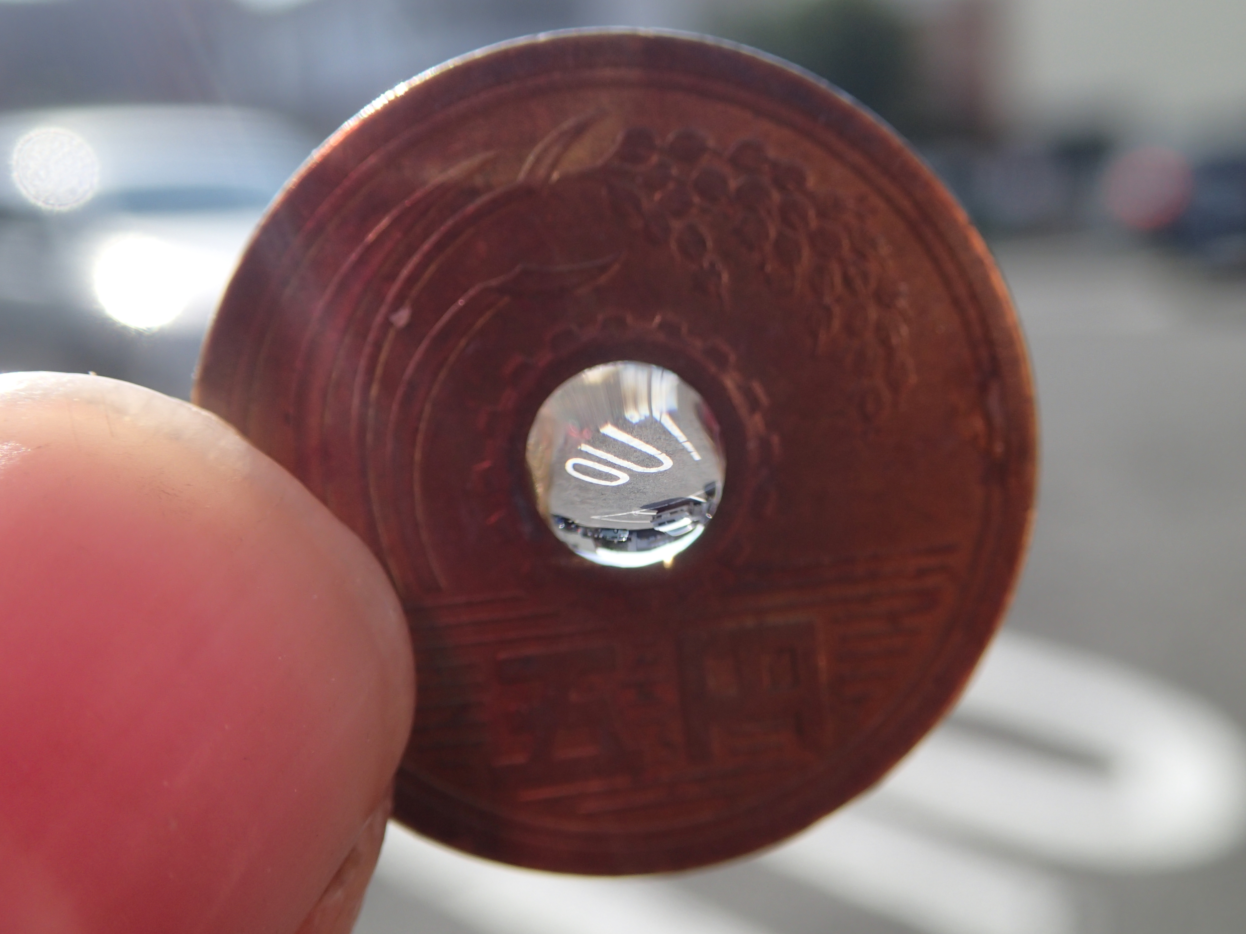 自宅近所で駐車場の「OUT」の文字を撮影。5円玉の背後にも同じ風景の一部がピンボケで写っている。