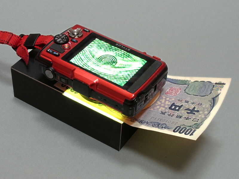 「OLYMPUS STYLUS TG-2 Tough」の強力なマクロ機能を活かした「透過光マクロ撮影ユニット」を制作してみた。光源にはiPhone 5のLEDライトを利用しているのがミソである。ご覧の通り、千円札の野口英世の目が画面いっぱいに写るほどの拡大率だ。透過光なので、印刷の裏が透けて面白い写真が撮れる。
