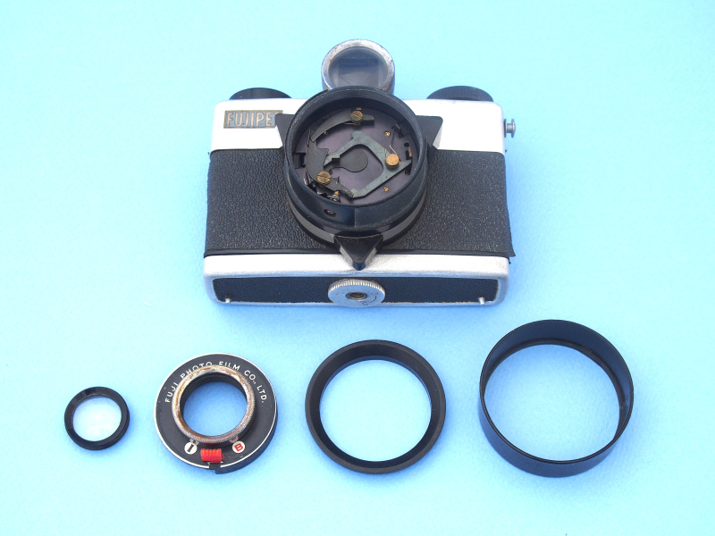 さて、工作するにはレンズの分解が必要だが、作業は先端にねじ込まれたリングを外すだけで簡単に行える。レンズ（手前左）は、1枚構成の単玉でガラス製である。単玉レンズは緒収差が補正しきれず、そのためフィルムガイドレールが湾曲した構造になっている（先の写真を参照）。またこれだけシンプルながら、引き出し式のレンズフード（手前右）が内蔵されている点にこだわりが感じられる