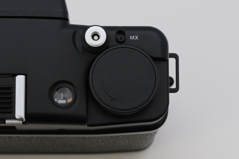 中央にあるのが、フィルム巻き上げダイヤル。左にあるのがフィルムカウンターで、撮影枚数が表示されます。その奥にある銀色のボタンがシャッターボタンです。シャッターボタンには、ケーブルレリーズ（付属）を装着できます。シャッターボタンの右にある「MX」は多重露光スイッチ。このレバーを下げると、同じコマで何度でもシャッターを切ることができます。