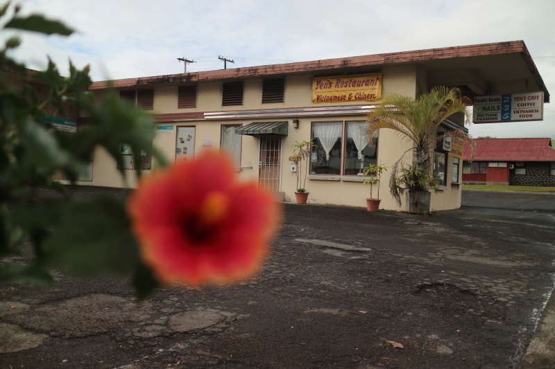 ピントを古いレストランに合わせる。手前にぼけたハイビスカスがあることで、この場所が南国であることが分かる。赤い色が差し色となった