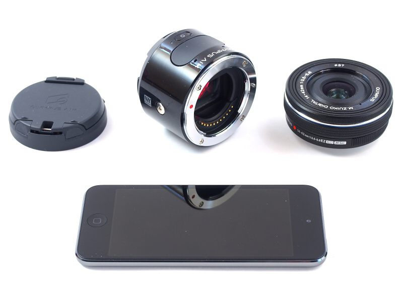 OLYMPUS AIR A01のカメラ本体（後中）は操作部として電源ボタン、シャッターボタン、レンズマウントのみを備える。構図確認を含むカメラ操作はWi-Fiで接続したスマートフォンによって行い、これを本体に装着するためのアダプター（後左）も付属している。レンズはマイクロフォーサーズマウントなら何でも装着できる。なお、今回は発売直前のOLYMPUS AIR A01をお借りしたため、スマートフォンのかわりにアプリがインストールされたiPod touch を使用する