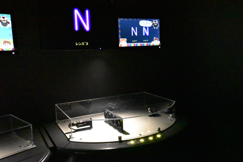 「レンズの実験室」コーナーのひとつ。手前のボタンを押すたびにレンズが切り替わり、「N」の字の周りのにじみが変化する。