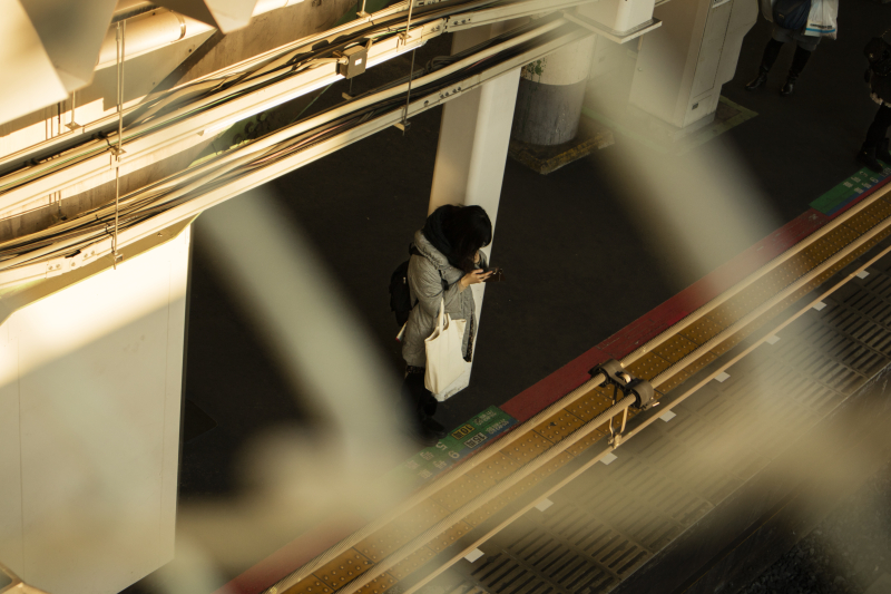 夕方の駅のホーム。線路を跨ぐ陸橋から撮影した。スマートフォンを見ながら電車を待つ女性を金網越しに狙い、そっとのぞいているような雰囲気を出している<br class="">キヤノンEOS M3／EF-M15-45mm F3.5-6.3 IS STM／45mm（72mm相当）／絞り優先AE（F6.3、1/250秒、-0.7EV）／ISO 200／WB：くもり