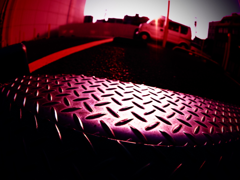 地面すれすれのローポジションで駐車場のプレートにピントを合わせた。プレートの凹凸模様がデフォルメされて、遠近感が強調された<br class="">オリンパスOM-D E-M10／M.ZUIKO DIGITAL ED 8mm F1.8 Fisheye PRO／8mm（16mm相当）／絞り優先AE（F2.8、1/4,000秒、-0.7EV）／ISO 200／WB：オート／アートフィルター：クロスプロセスII