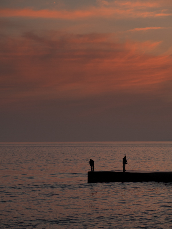 夕焼けの中に佇む釣り人を望遠レンズで切り取った1枚。日が沈んだ直後ならではの美しいグラデーションが2人のシルエットをより際立たせている。<br class=""><span class="fnt-85">オリンパス OM-D E-M1 / M.ZUIKO DIGITAL ED 75mm F1.8 / 75mm（150mm相当） / 絞り優先AE（F2.8、1/160秒、-0.7EV） / ISO 200</span>