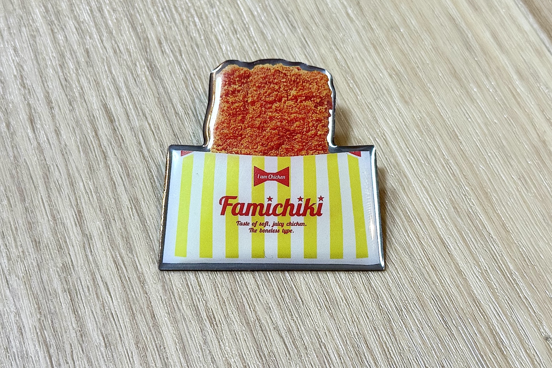 ファミチキ ピンバッジ。袋を半分だけ切って手を汚さずに食べられるあの見慣れたスタイルがそのまま再現されている