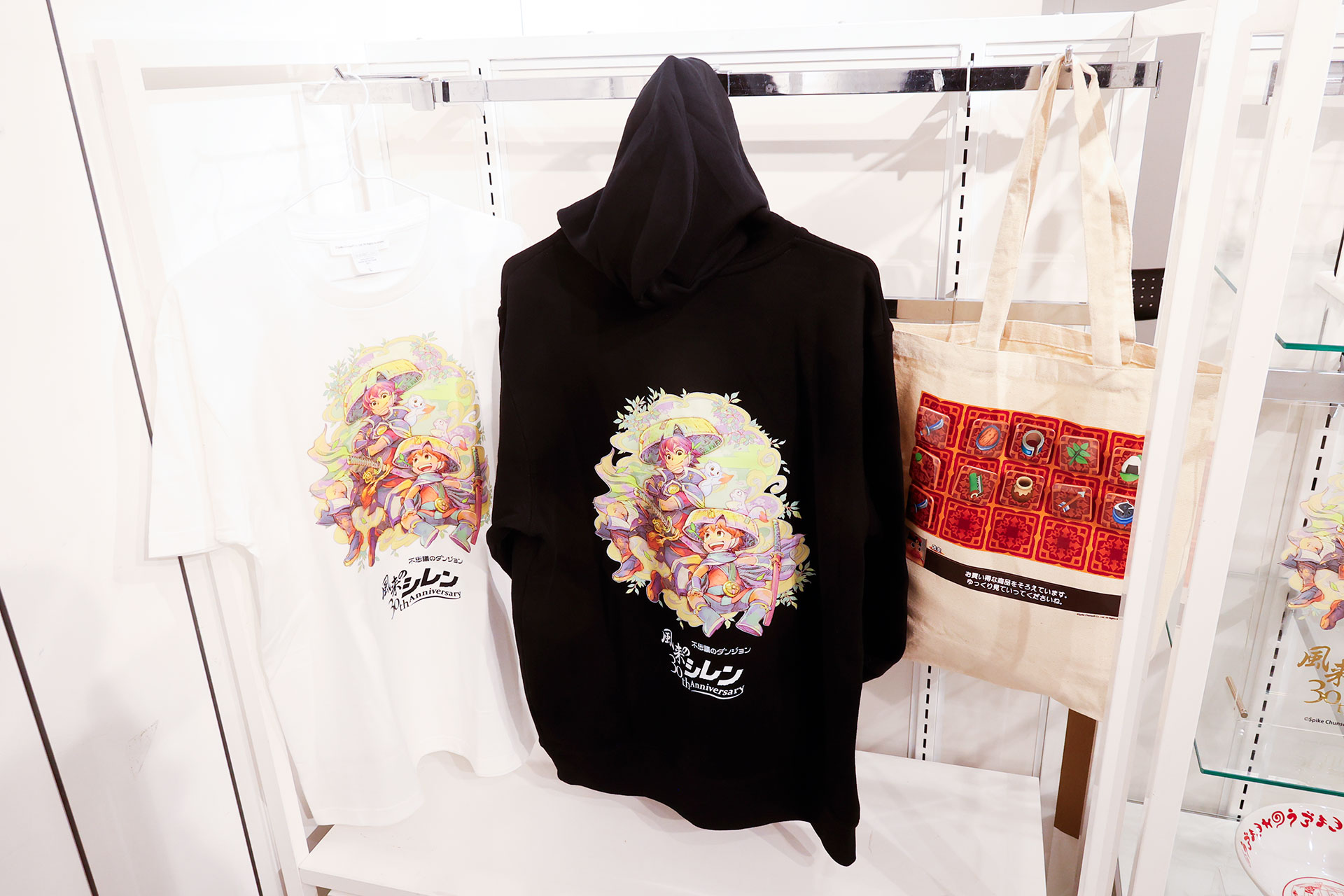 こちらが長谷川薫氏の描き下ろしイラストが描かれたTシャツとパーカーだ。トートバッグには写真に写っているようなバッジもひとつランダムで付属している