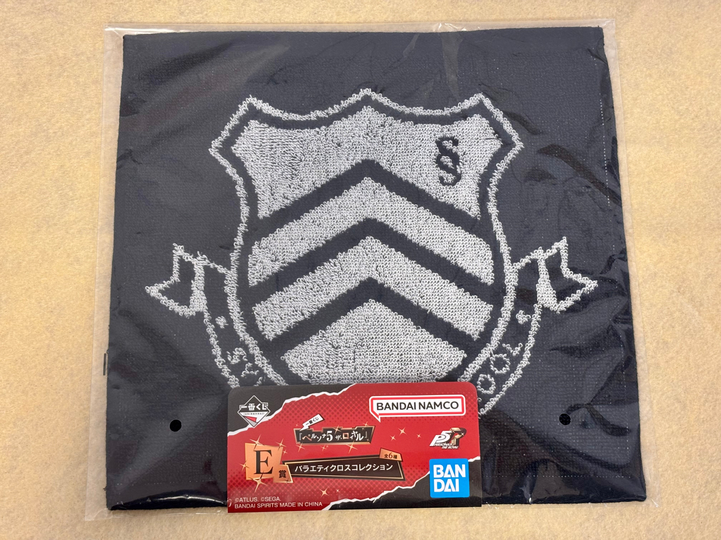 秀尽学園エンブレム：黒地に白のステッチ風デザインで校章があしらわれている。普段使いしやすく、さりげないファンアピールに最適