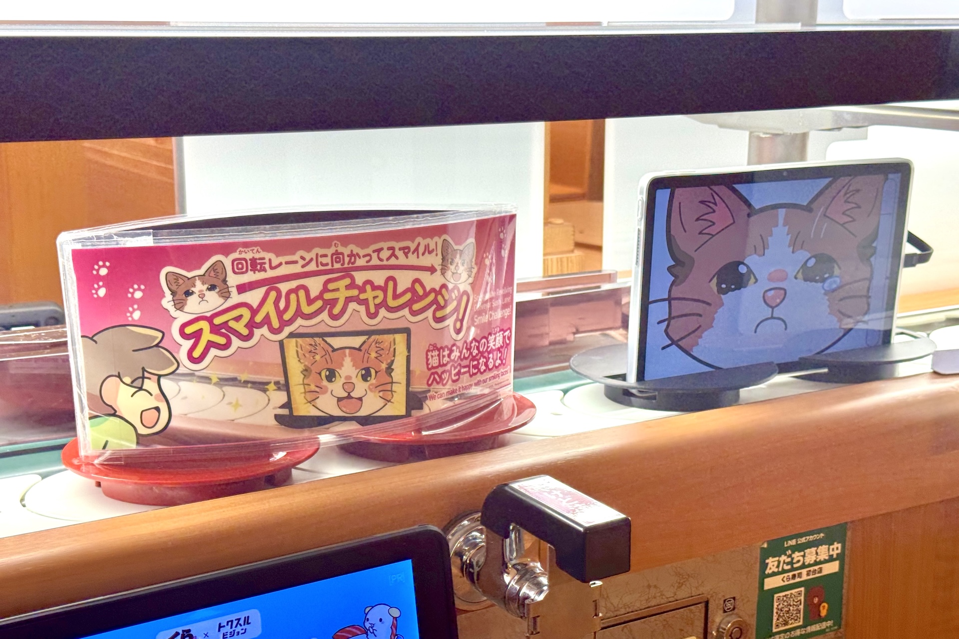 くら寿司では回転レーン×AI活用のサービス「スマイルチャレンジ」を2025年9月より導入中。回転レーンに流れてくる泣いている猫が映るモニターに向かって笑顔を見せると、AIがその笑顔を判定し、その結果に合わせて猫の表情が変化するというものだ