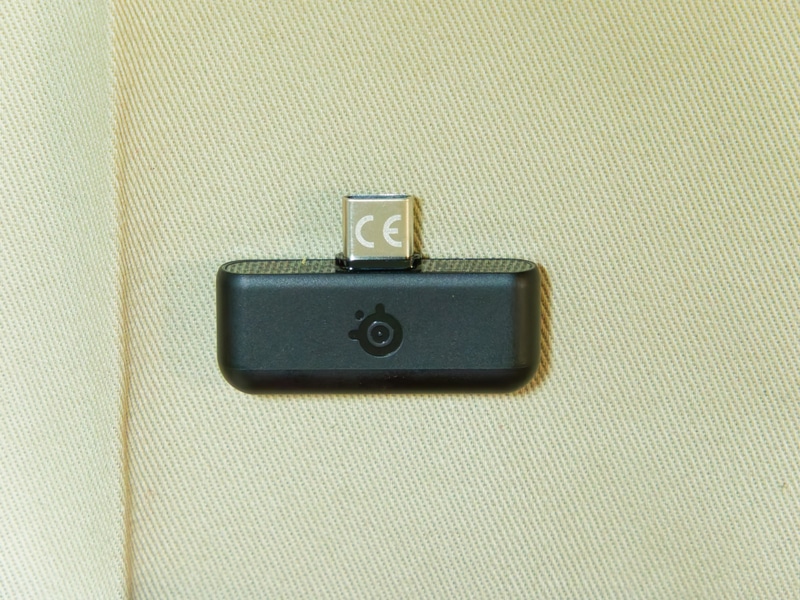 Arctis Nova 7 Gen 2のUSB-Cドングル。ドングルと本体はあらかじめペアリング設定がされた状態で出荷されている。改めてペアリングを行う場合は、ドングルを再生機器のUSBポートと接続したうえで、ドングルのペアリングボタンを長押し、さらにヘッドセット側の電源を一度切ってから電源ボタンを長押ししてこちらもペアリングモードにすることでかんたんにペアリングが実行できる