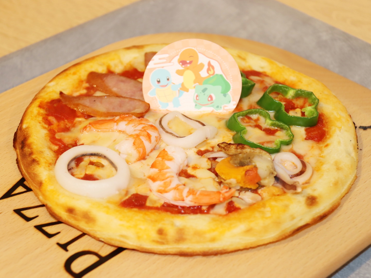 フシギダネ＆ヒトカゲ＆ゼニガメの三位一体ピザ 2,200円/ピザの上にピーマン、ソーセージ、魚介類が乗ったピザ。3種の具材でそれぞれフシギダネ、ヒトガケ、ゼニガメを表現している