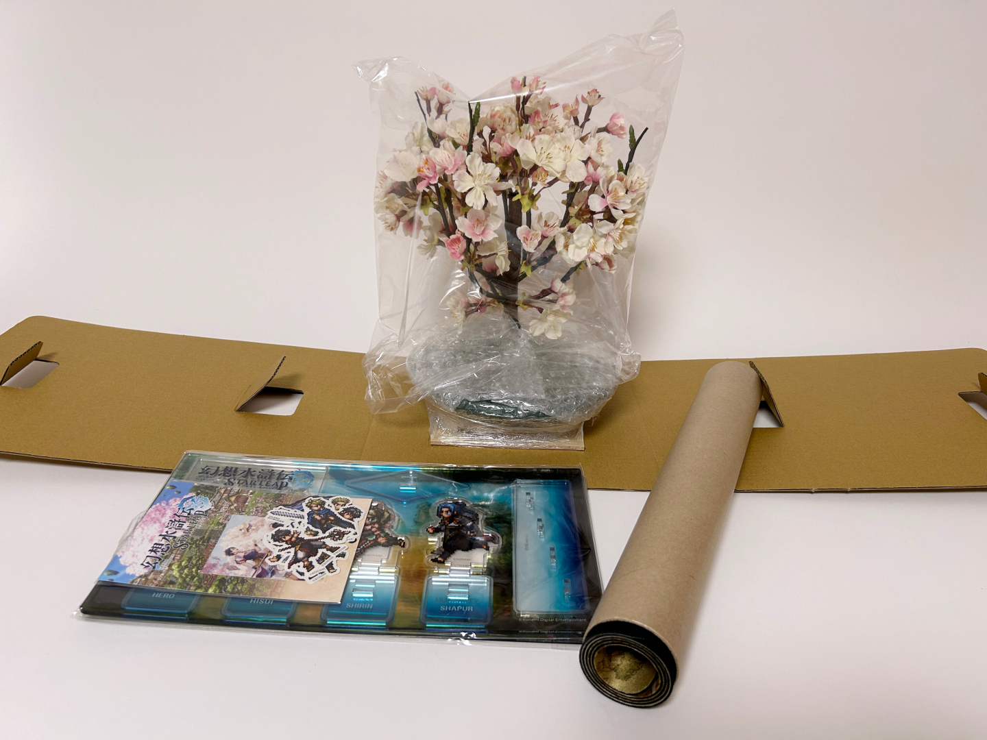 中身はこのような感じで収納されている。取り出して並べてみたが、奥に見えるのが桜盆栽で手前右側がデスクマット、左がアクリルスタンドとステッカーのセットとなる