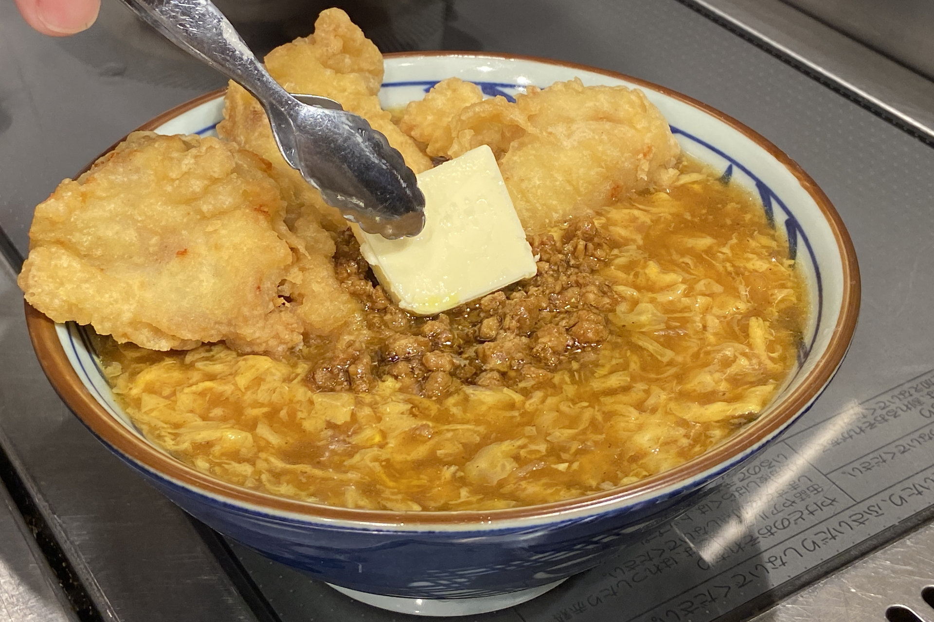 京風のカレーあんかけをベンチマークにしたカレーうどんの上に、塩胡椒とカレー粉で味つけした鶏天を3つ、カレー味のそぼろ、無塩バターをトッピング