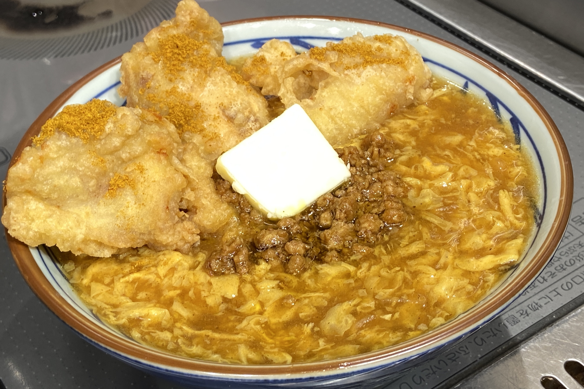 京風のカレーあんかけをベンチマークにしたカレーうどんの上に、塩胡椒とカレー粉で味つけした鶏天を3つ、カレー味のそぼろ、無塩バターをトッピング