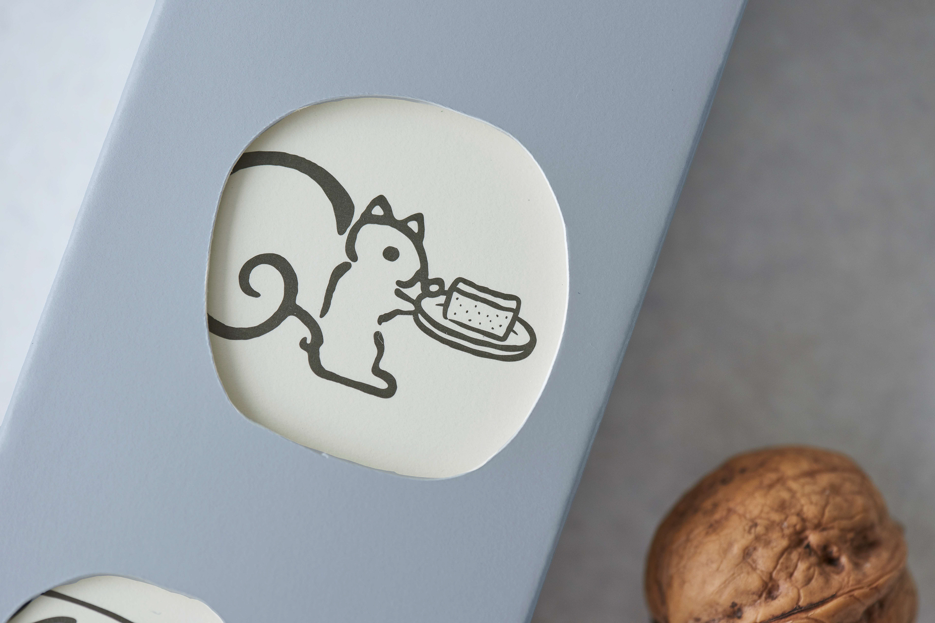 お皿にのせたチーズケーキを運ぶ鎌倉紅谷のシンボルキャラクター「リスくん」と、Mr. CHEESECAKEのキャラクター「ミスチーくん」と「子リスくん」の出会いをパッケージにデザイン