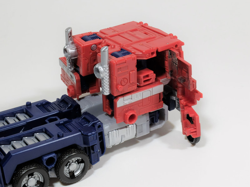 ここで足を90度倒して、腕部を本体に収納する。腕部の収納スペースはここしかない、というくらい明確に収納可能なスペースがキッチリ空いているので、そこに腕の形状を合わせて収納する。あとはトラクターヘッドのフロント部の扉を全て閉じる。全てがピタッと収まるようになっているので、ピッタリ合わない場合は、状態を再度確認しよう。あとは太腿部の部品を展開すればトランスフォームが完了となる。