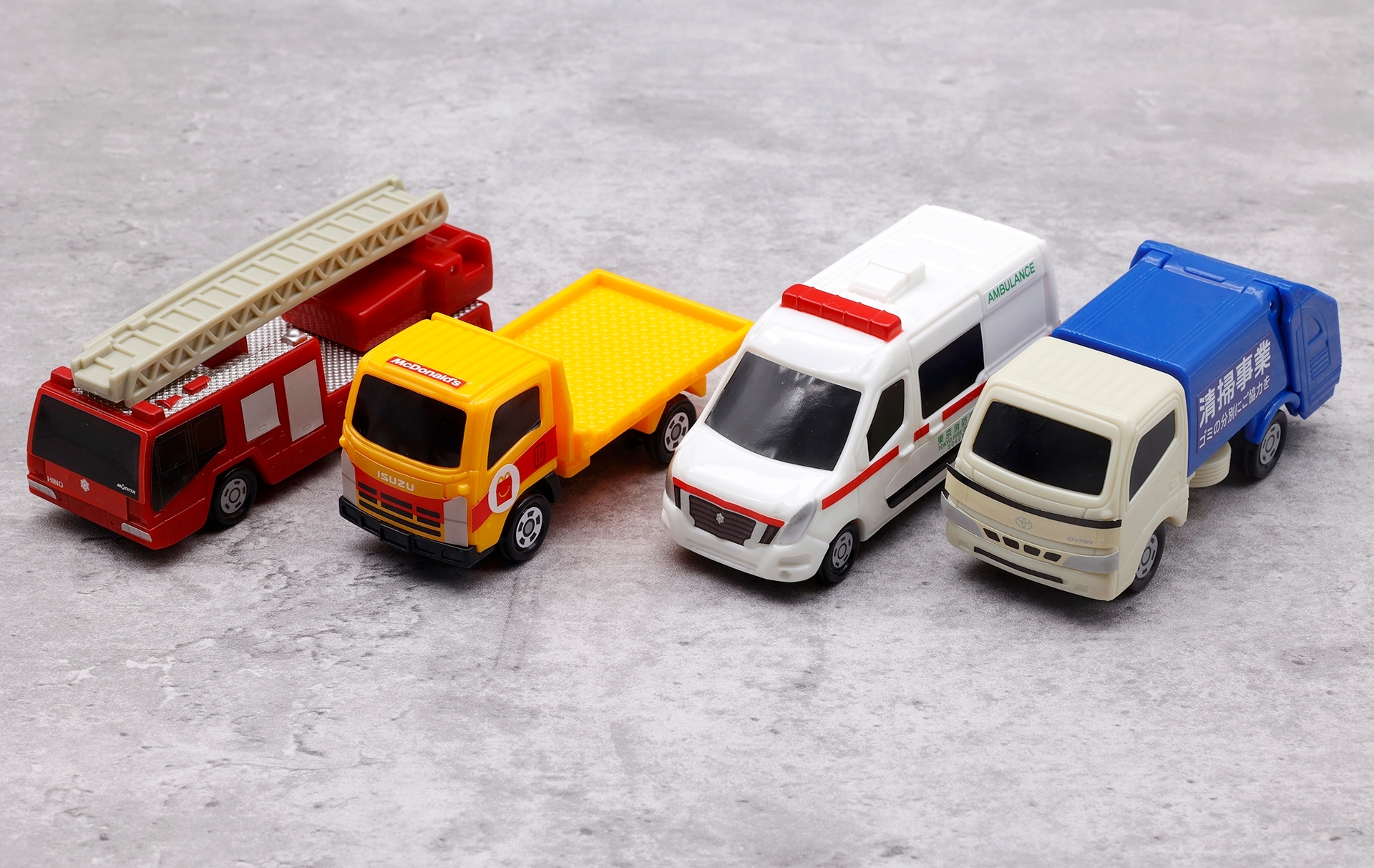 こちらが第1弾のハッピーセット「トミカ」に登場するミニカー。写真左から「日野はしご付消防車（モリタ・スーパージャイロラダー）」、「いすゞ エルフ 車両運搬車」、「日産 NV400 EV 救急車」、「トヨタ ダイナ 清掃車」