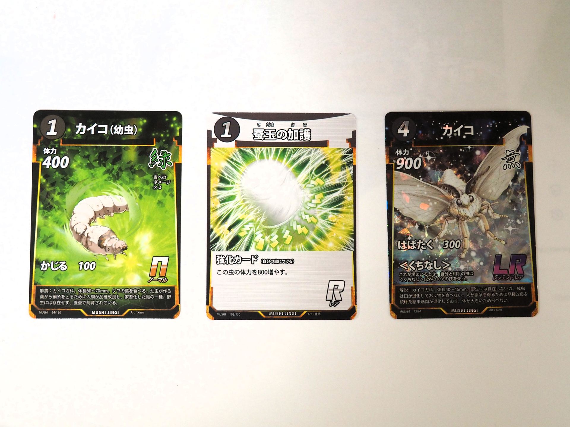 蟲神器には第5弾までの時点で3枚のカイコ関連カードが登場している。左から第1弾の緑1コストNカード「カイコ（幼虫）」、第1弾の1コストR強化カード「蚕玉の加護」、第4弾の無色4コストLRカード「カイコ」