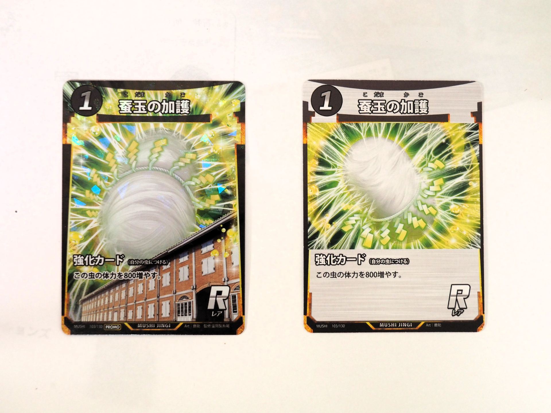 左がイベント限定プロモカードの「蚕玉の加護」。右が通常の「蚕玉の加護」。限定プロモカードはイラストが違うだけでなく、キラ加工もされている。背景に富岡製糸場の西置繭所が描かれているのもポイントだ