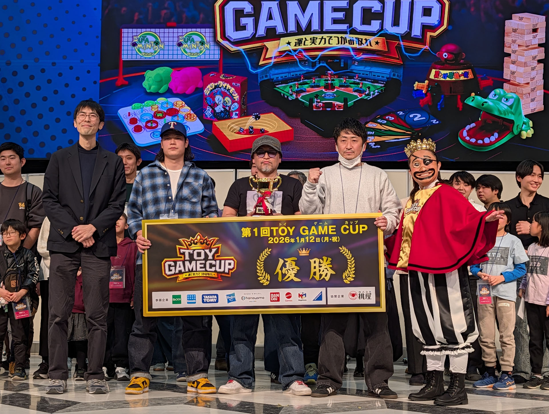 優勝した「ひしんたい」のメンバーとキンタロー氏、高橋氏を囲んでのフォトセッションより。高橋氏は優勝した「ひしんたい」について、「ブタミントン」は強そうな見た目だったが、他のゲームでも強かったことが優勝に繋がった。見た目でトイゲームが好きそうには見えないのに、こういう大会に出てくれるなど、改めてトイゲームの間口の広さを感じたとコメントした。また、キンタロー氏は「今回もリアルに人が集まったことでこんなに素晴らしいドラマが生まれた。改めて優勝した「ひしんたい」のみなさん、おめでとうございますとコメントした