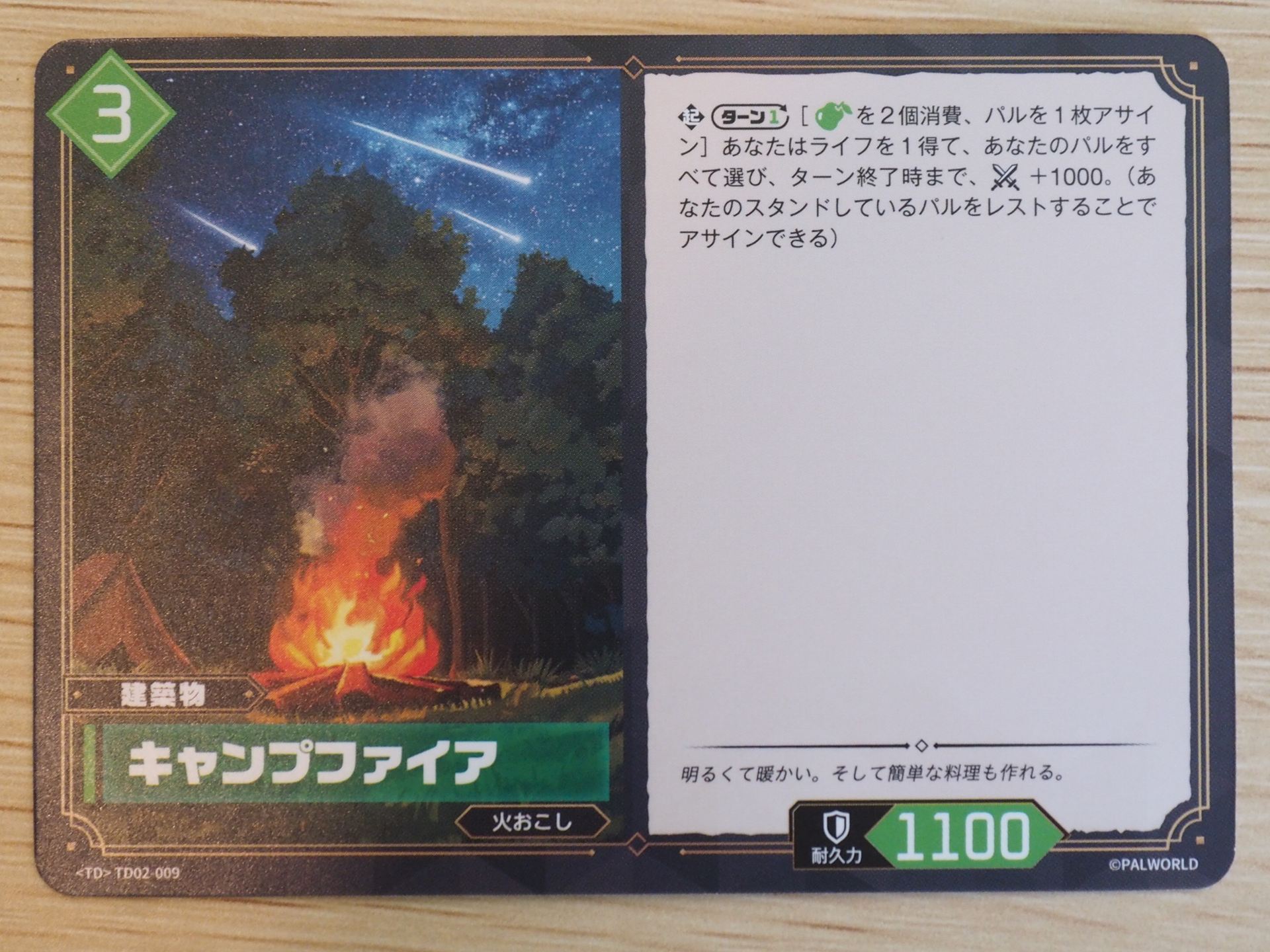 緑3コストの「建築物カード: キャンプファイア」。ターンに1回、『食材』を2個消費してパルを1枚アサインすることで、ライフを1得て、あなたのパルをすべてターン終了時まで戦闘力を＋1000することができる