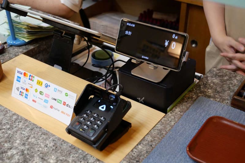 新しい運用スタイル。レシートプリンター上にカスタマーディスプレイ用スマートフォンを設置して、その前のカウンターにM010を固定したmC-Standを設置した