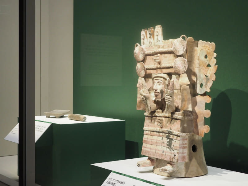 写真手前は、1325～1521年に作られた熟したトウモロコシの女神をかたどった《チコメコアトル神の火鉢(複製)》。原品はメキシコシティのトラワク地区で出土し、メキシコ国立人類学博物館蔵。写真奥が、250～550年頃のテオティワカン文明で使われていた、トウモロコシをすりつぶすための《メタテ(石皿)》と《マノ(石棒)》。テオティワカン考古学ゾーン蔵