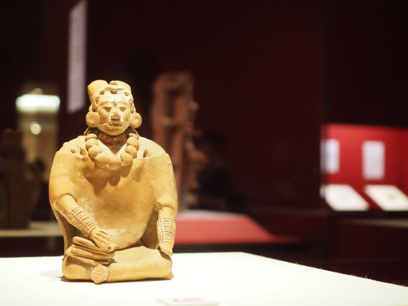 《書記とみられる女性の土偶》も、600～950年頃に制作されたハイナの出土品。大きな首飾りや腕輪、右手には書物が見られる。メキシコ国立人類学博物館蔵。背後にあるのは《香炉台》