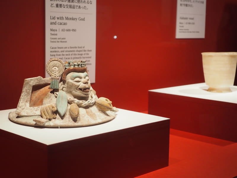 手前は、600～950年に作られ、トニナで出土したトニナ遺跡博物館蔵の《猿の神とカカオの土器蓋》。首に猿の好物であるカカオの実の装飾がある。右はユカタン州で出土した800～1530年制作の《アラバスター容器》。炭酸カルシウムが層状に堆積した半透明のアラバスターで作られたという。メリダ マヤ世界大博物館蔵