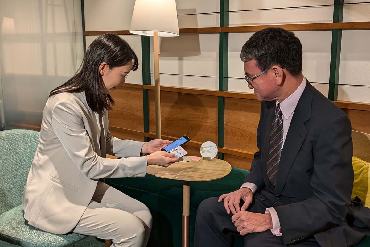 2024年8月に三井住友銀行の支店で当時のデジタル大臣の河野太郎氏が自身のマイナンバーカードを提示して本人確認の実証実験に参加しているところ。スマートフォン搭載のマイナンバーカードでは直接券面情報が確認できないため、このような「マイナンバーカード対面確認アプリ」が必要となる。もちろん、ICチップを読むのでセキュリティ対策の意味合いもある