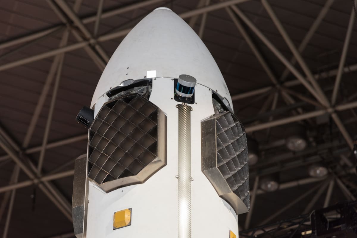 HONDAの再使用ロケット上部。着陸時の姿勢を制御する、うちわ状の制御翼(グリッドフィン)が4面に取り付けられています。制御翼の上部には、姿勢や高度を計測するセンサー類。加速度・ジャイロセンサー、LiDAR、GPSアンテナを備えているとのこと。円筒状の2種類のセンサーのうち、青いテープを撒いたようなセンサーは米Velodyne社のLiDARではないかとみられています(撮影:小林伸)