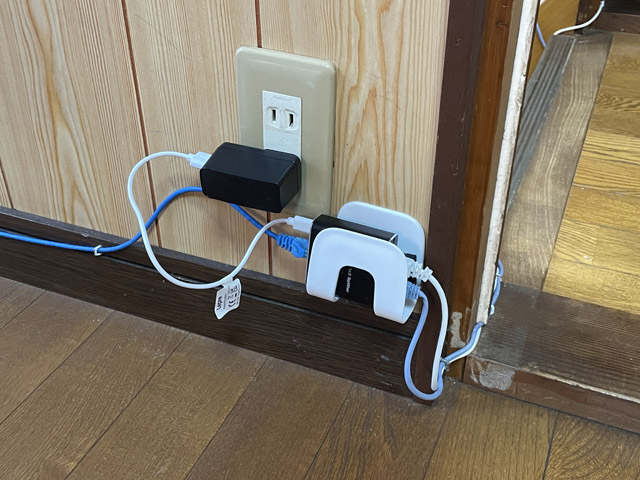 仕事部屋のドア付近に3ポートのスイッチングハブを設置。左側の青いLANケーブルはWi-Fiルーターへ。右側の水色のLANケーブルは1階へ。白のLANケーブルはオーディオ部屋へ。電源はUSBアダプター（Type-A出力）から、手元にあった一番短いケーブルでスイッチングハブに給電。ACアダプターよりスマートに設置できる