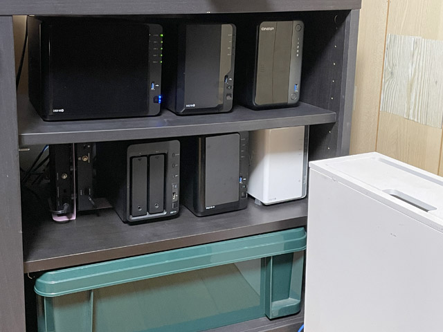 6台のNAS。上段右端の1台がQNAP。残りの5台はSynology。中段の左端は2.5Gbps×8ポートのスイッチングハブが2台。下段の緑の箱は、コンテナボックスを加工した自作の大型ケーブルボックス。右の白いケーブルボックスにONU、Wi-Fiルーターが入っている