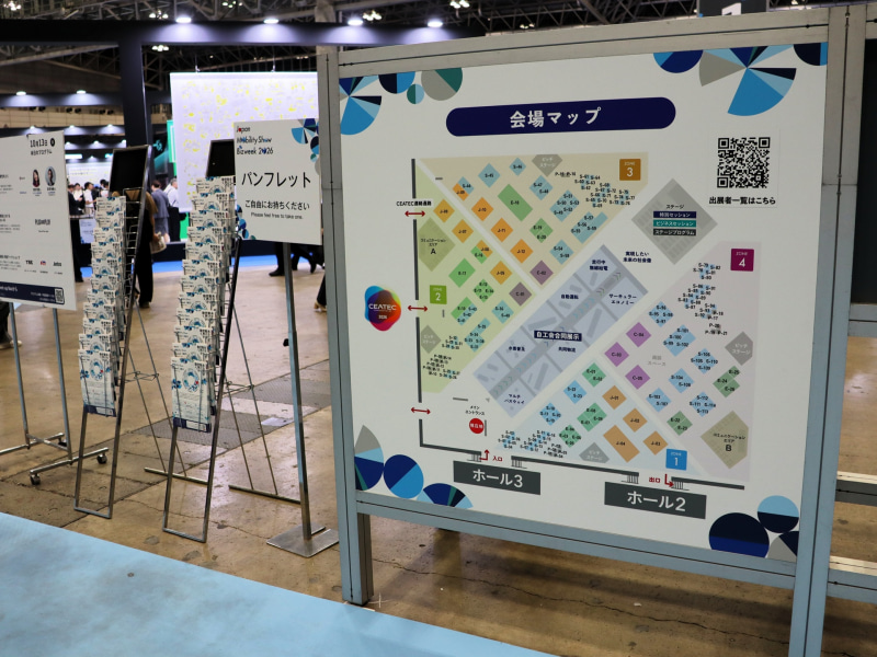 「Japan Mobility Show Bizweek 2026」の展示ホールに降りてすぐのところに会場マップが設置されている。紙の会場マップは、その横に置いてある「パンフレット」の内面に掲載されている